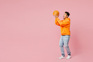 Full body side view young man of African American ethnicity fan in yellow hoody casual clothes cheer up support basketball sport team hold ball watch tv live stream isolated on plain pink background