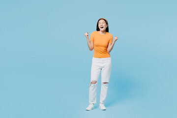 Full body young woman of Asian ethnicity she wear orange t-shirt casual clothes doing winner gesture clench fist isolated on plain pastel light blue cyan background studio portrait. Lifestyle concept.