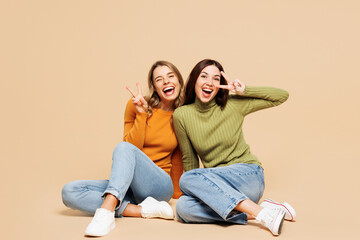 Full body young friend two women wear orange green shirt casual clothes together sit looking camera cuddle hug embrace show cover eye with v-sign isolated on plain beige background. Lifestyle concept.