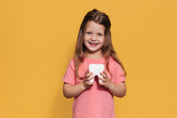 A smiling girl with healthy teeth holds a plastic tooth in her hands on a yellow insulated background. A place for your text. Oral hygiene. Pediatric dentistry. Prosthetics. Rules for brushing teeth.