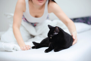 A young woman in a white T-shirt is trying to move and shoo her black cat out of her white bed - 742356760