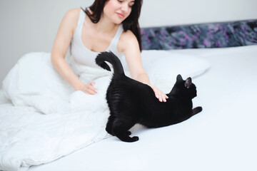 Young beautiful woman in a white tank top petting her black cat on her white bed - 742356770