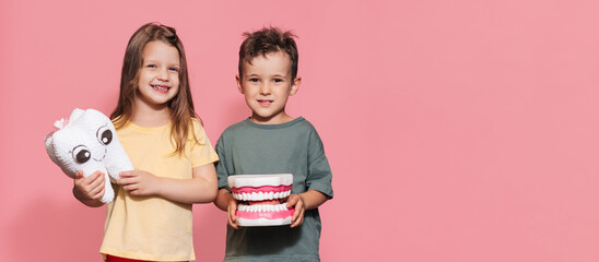 A smiling boy and a girl with healthy teeth hold a toy tooth in their hands on an isolated background. A place for your text. Oral hygiene. Pediatric dentistry. Prosthetics. Rules for brushing teeth. - Powered by Adobe