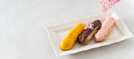 Delicious eclairs banner. Set of eclairs on a white plate on a light background with hyacinth flower. Copy space, side view. Traditional French dessert recipe. Confectionery, cafe web line