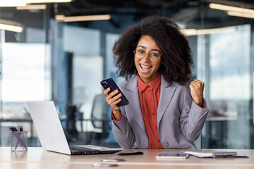 Cheerful african american female in business suit with mobile gadget in hand making victory gesture...