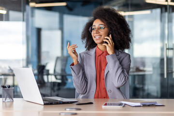 Obraz premium Happy latino entrepreneur using mobile phone for connecting while sitting by desktop with open laptop. Young busy executive making pleasant call while looking at window in corporate office building