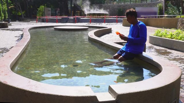 A man watching mobile during the hot spring bath in the seat of hot spring pool.Seat in the hot spring pool for sitting. .Tourists soak their feet in the hot spring pools to relax.