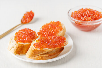 Sliced loaf of bread or canape with butter spread and salted red caviar or salmon fish roe served on plate white wooden table with glass bowl and spoon as healthy appetizer full of protein and omega 3