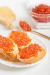 Plate with tree slices of bread or canape with butter and salted red caviar or salmon fish roe served on white wooden table with glass bowl and spoon as appetizer for holiday celebration dinner