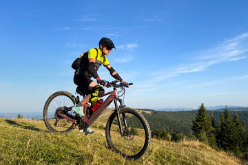Obraz premium Cyclist man riding electric mountain bike outdoors. Male tourist biking along grassy trail in the mountains, wearing helmet and backpack. Concept of sport, active leisure and nature.