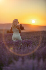 Blonde woman poses in lavender field at sunset. Happy woman in white dress holds lavender bouquet....