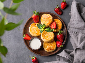 Cottage cheese, cheesecakes or syrniki with sour cream, strawberries and mint leaves on a wooden plate on a dark background with napkin and branch.