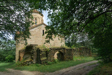 Fototapeta na wymiar Ancient church in the forest. Bricks and stone wall. Partially destroyed arched extension at the entrance. Orange tiled roof. Grass lawn, traces of an earthen road.