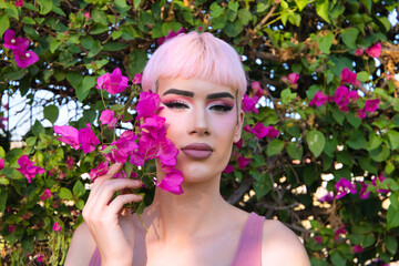 Portrait of young gay boy with pink hair and make-up has next to his face a purple bougainvillea...