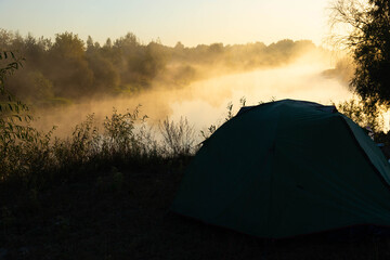 Obraz premium Green tourist tent by the river at sunrise, with morning autumn fog on the water. Outdoor tourist landscape.