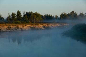 Foggy morning on a European river with fresh green grass in the sun. The rays of the sun through the tree.
