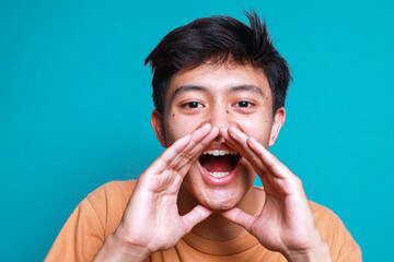 Young Asian Man Shouting Over Blue Background. Shouting with Hands Cupped Around Mouth.  