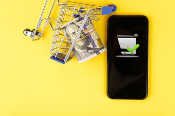 Online shopping, telephone and shopper trolley concept with roll of one hundred dollar bills on yellow background. Copy space.