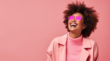 A happy laughing African American girl with an Afro hairstyle, wearing sunglasses, a sweater, a leather jacket on a pink background with a copy space. Positive Emotions, Fashionable Youth Clothes.