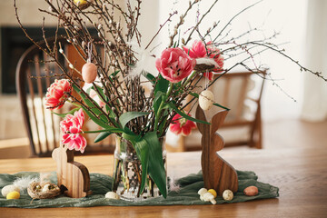 Stylish tulips bouquet with willow branches, easter eggs and wooden bunny figurines decor on wooden...