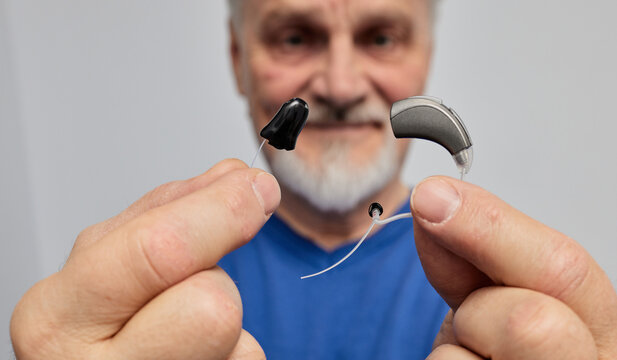Senior man with hearing problems chooses between behind-the-ear hearing aid and in-the-ear hearing aid by holding them in front of her. Hearing aids and solutions