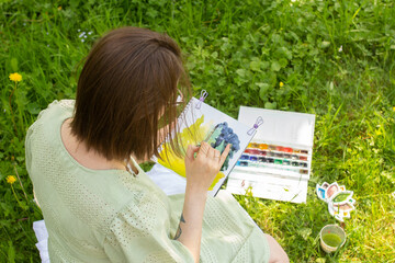Female artist painting still life on plein air in park,. Woman drawing watercolor sketch and sitting on grass outdoor