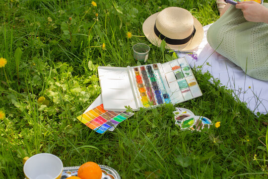 Watercolor palette laying on green grass near woman drawing and painting on plein air in the summer park on sunny day