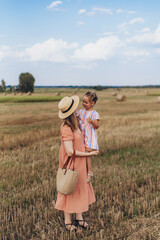 A young mother in a long dress and a straw hat leads a little daughter by the hand. Both walk across a field of harvested wheat. Against the background of a bale of straw