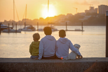 Children, boys, brothers, enjoying sunset over river with their pet maltese dog and mom, boats, sun, river - obrazy, fototapety, plakaty