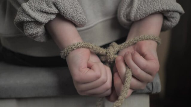Close-up of a child's small hands tied behind his back with a rope. The child is trying to free his hands from the shackles of the rope. family violence against children. Child in captivity, hostage