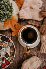 Big mug of hot dark coffee, sweet candies, autumn leaves and handmade woolen clothes on wooden background, vintage scene