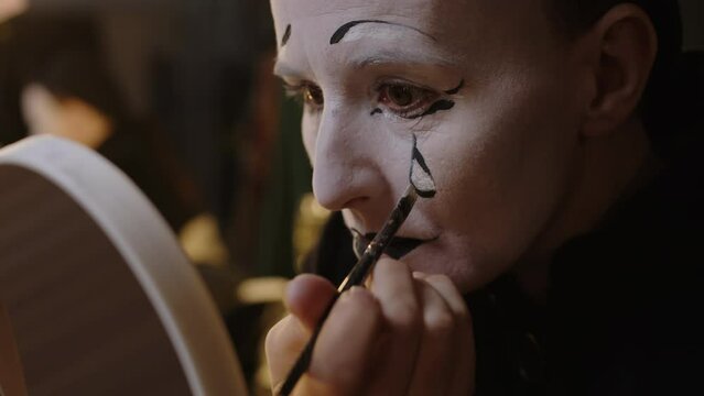 Medium closeup of adult mime actress drawing black tear on her face while doing sad makeup, preparing for stage act in backstage room