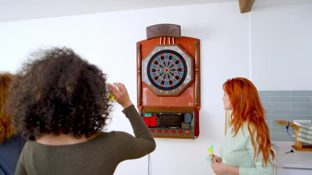 Two women playing darts in a kitchen, one aiming while the other waits with darts in hand. 