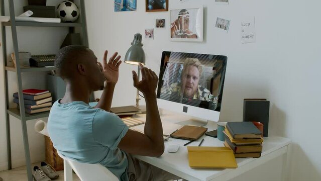 Slow motion shot footage of modern African American student sitting at desk in front of computer monitor discussing university project with groupmate on video call