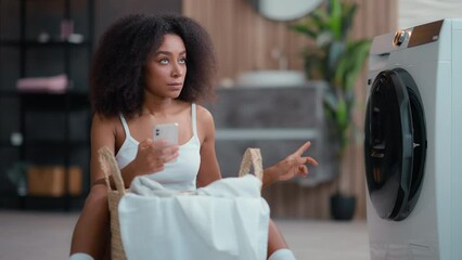 African American woman ethnic girl housewife do housework housekeeping basket with cloth dirty clothes reading instruction using washing machine laundry appliance chatting mobile phone smartphone hold - Powered by Adobe