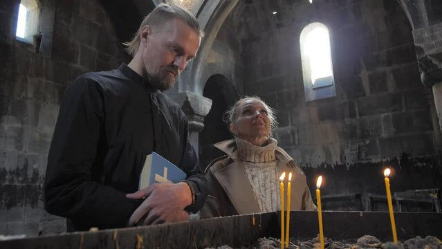 A young attractive priest holding a bible standing with a pretty woman at the altar with burning candles and silently looking at the fire of the candles. the role of religion in people's lives