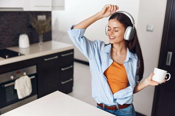 Fototapeta na wymiar Woman enjoying music with headphones and coffee in kitchen at home in morning sunlight