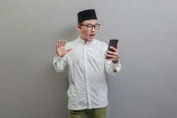 Portrait of an Asian Muslim man wearing a koko shirt and peci with shades of the fasting month, shocked while raising his hand while looking at his smartphone, isolated on a white background