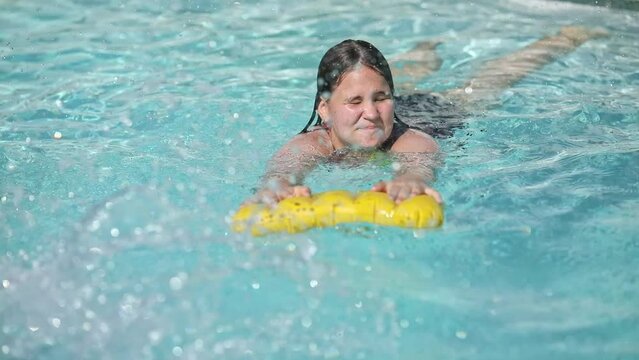 Happy teenager girl with yellow inflatable pillow in swimming pool with blue water on warm summer day on tropical vacations. Summertime activities concept.