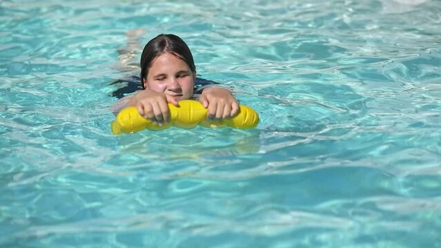 Happy teenager girl with yellow inflatable pillow in swimming pool with blue water on warm summer day on tropical vacations. Summertime activities concept.