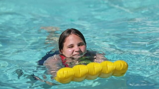 Happy teenager girl with yellow inflatable pillow in swimming pool with blue water on warm summer day on tropical vacations. Summertime activities concept.