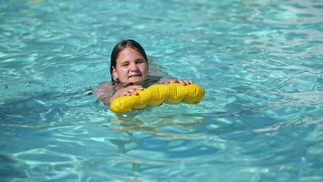 Happy teenager girl with yellow inflatable pillow in swimming pool with blue water on warm summer day on tropical vacations. Summertime activities concept.