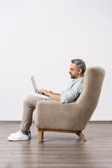 Side view of handsome man sitting in armchair and using laptop. White studio background.