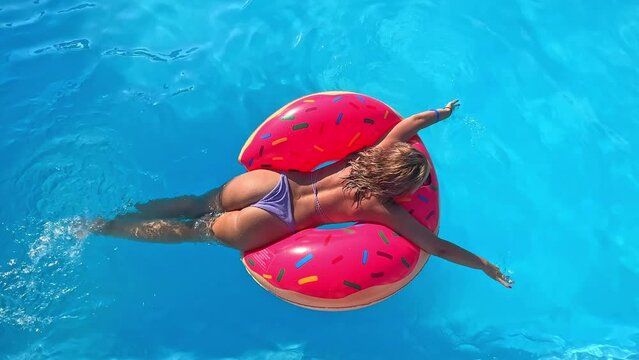 Young millennial woman girl in sprinkled doughnut float at swimming pool, has fun during festival, hotel, beach, event smiling with sunglasses on during summer. Vacation concept. Top view