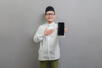 Portrait of an Asian Muslim man wearing a koko shirt and peci with shades of the fasting month, while stroking his chest and showing his smartphone, isolated on a white background