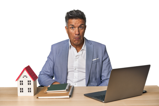 Middle-aged Hispanic businessman with real estate theme shrugs shoulders and open eyes confused.