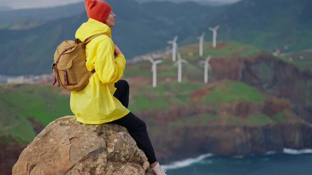 Attractive traveling woman in a yellow jacket and backpack looking at windmills in field 