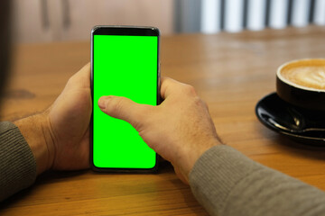 Young man sitting at cafe holding smartphone green mock-up screen in hand. Male person using chroma...