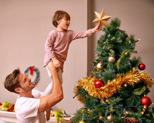 Father, son and happy with bonding by christmas tree for celebration and playing with star in the morning. Family, man and child with smile, relax and enjoying holiday season with decoration in home