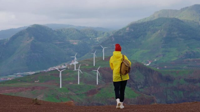 Attractive traveling woman in a yellow jacket and backpack looking at windmills in field 